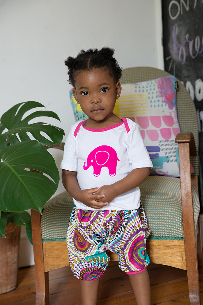 Child wearing a white shirt with a pink elephant design and colorful shorts, sitting on a chair.