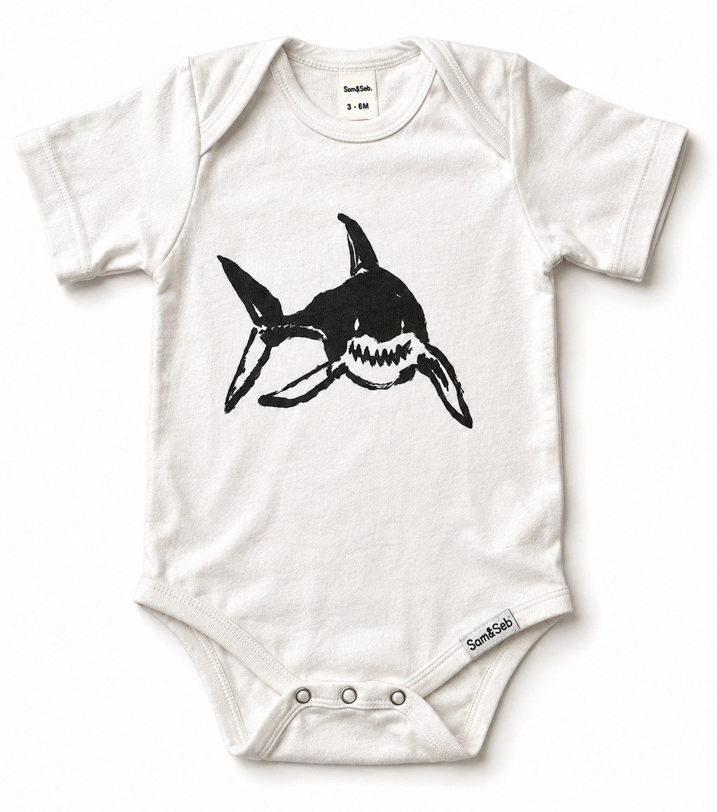 A white short-sleeve baby bodysuit featuring a black shark graphic on the chest, with snap closures at the crotch. The brand 'Sam & Seb' is visible on the inner neck label and the lower right hem.