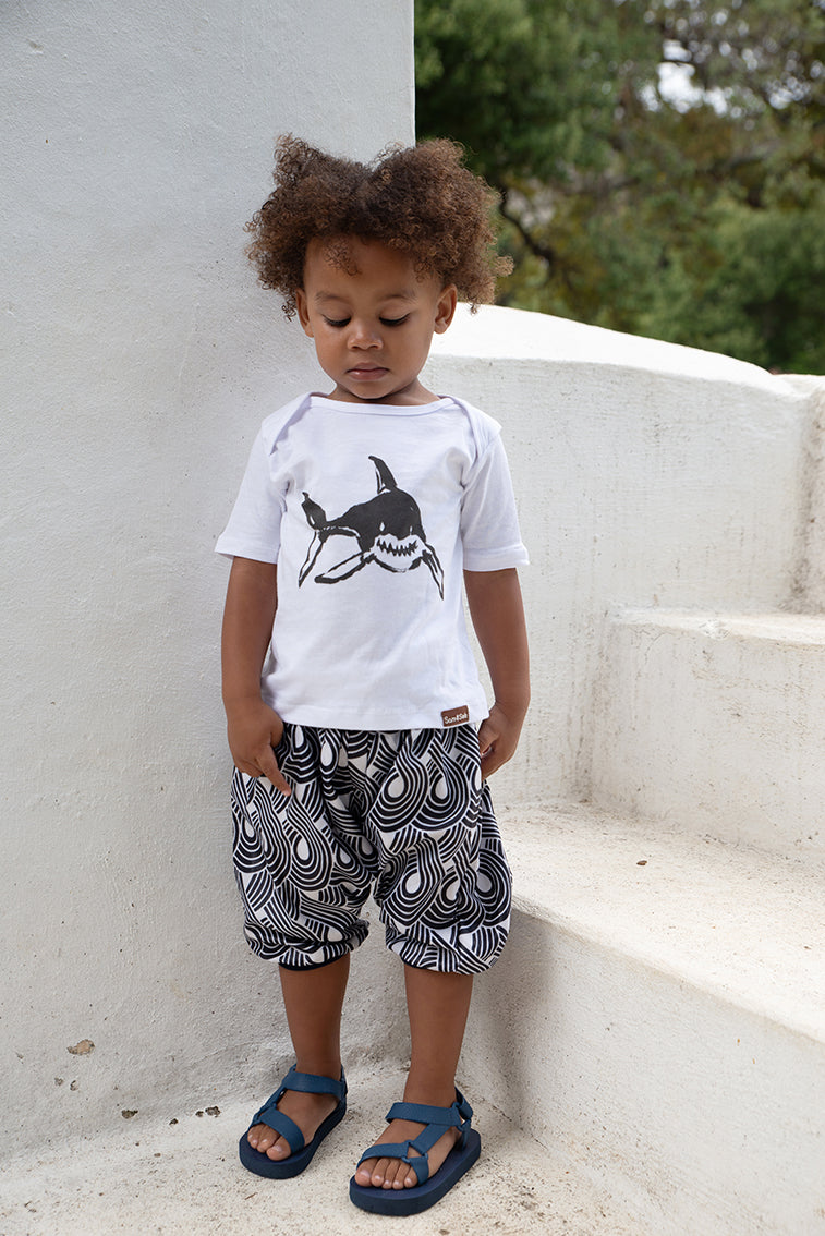 Child wearing a white t-shirt with a shark design and patterned shorts standing on a concrete surface.